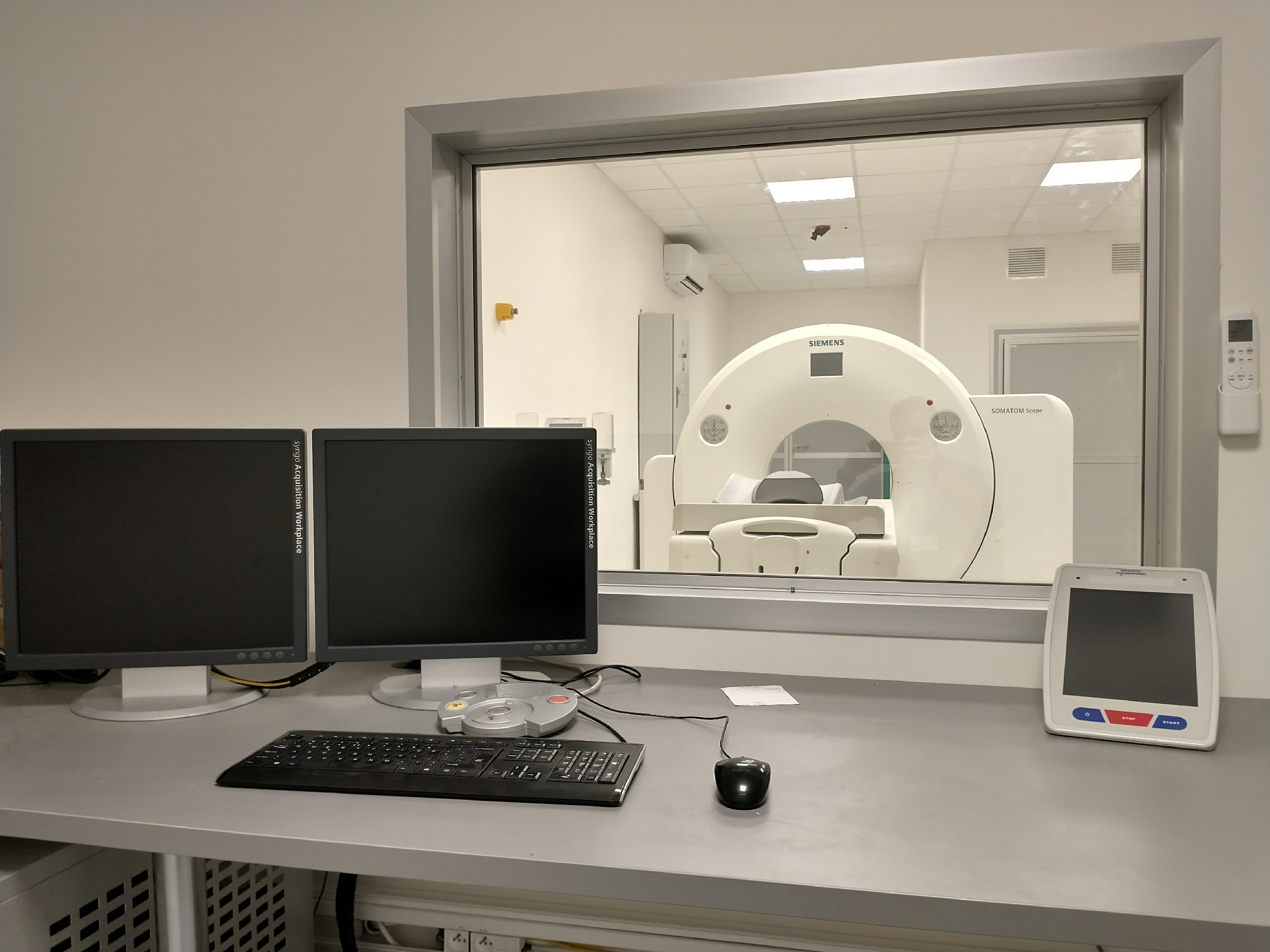 na obrazu widać dwa monitory stojące koło siebie wraz z klawiatura i myszą koloru czarnego, za oknem widoczne urządzenia do tomografii komputerowej. 